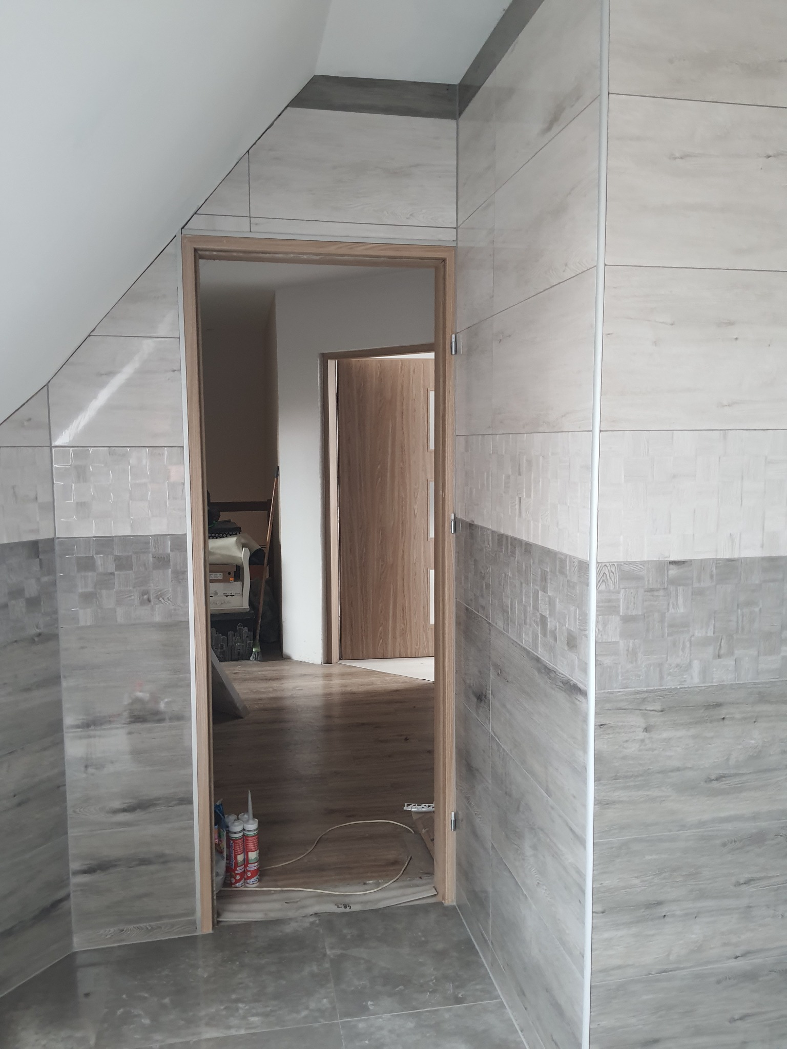 Wnętrze łazienki w trakcie wykańczania, widoczne płytki imitujące drewno na ścianach i podłodze, otwarte drzwi z widokiem na kolejne pomieszczenie, oraz tuby silikonu w rogu.