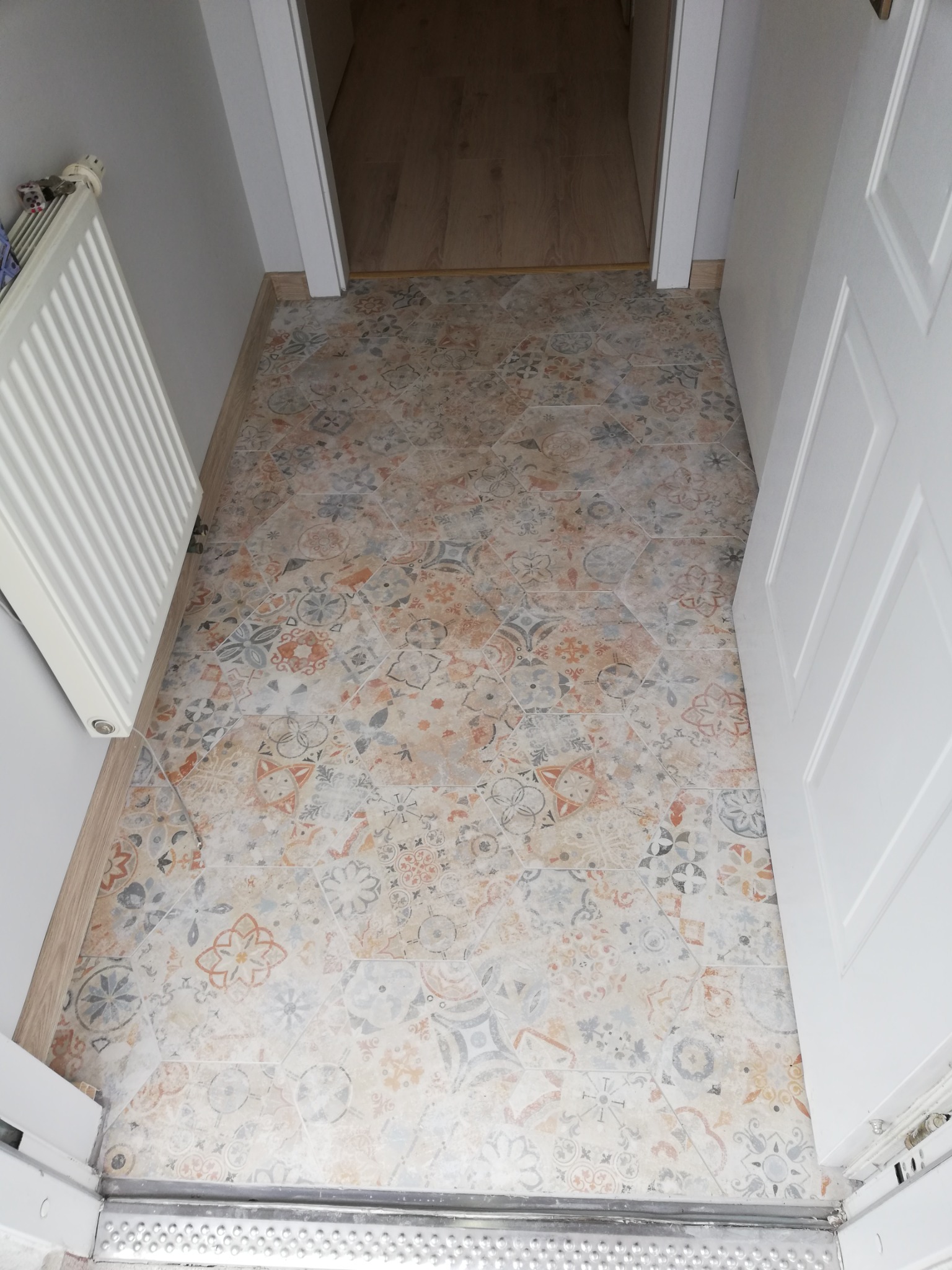 Podłoga wyłożona wzorzystymi, heksagonalnymi płytkami w odcieniach beżu, szarości i pomarańczy, widoczna przy białym grzejniku i otwartych białych drzwiach wewnętrznych.