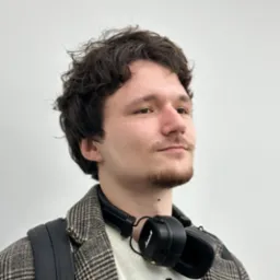 Młody mężczyzna z kręconymi włosami i delikatnym zarostem, po wizycie, spogląda w dal. Na szyi ma słuchawki Marshall, ubrany w sweter w zygzaki i marynarkę w pepitkę, z plecakiem na ramieniu.