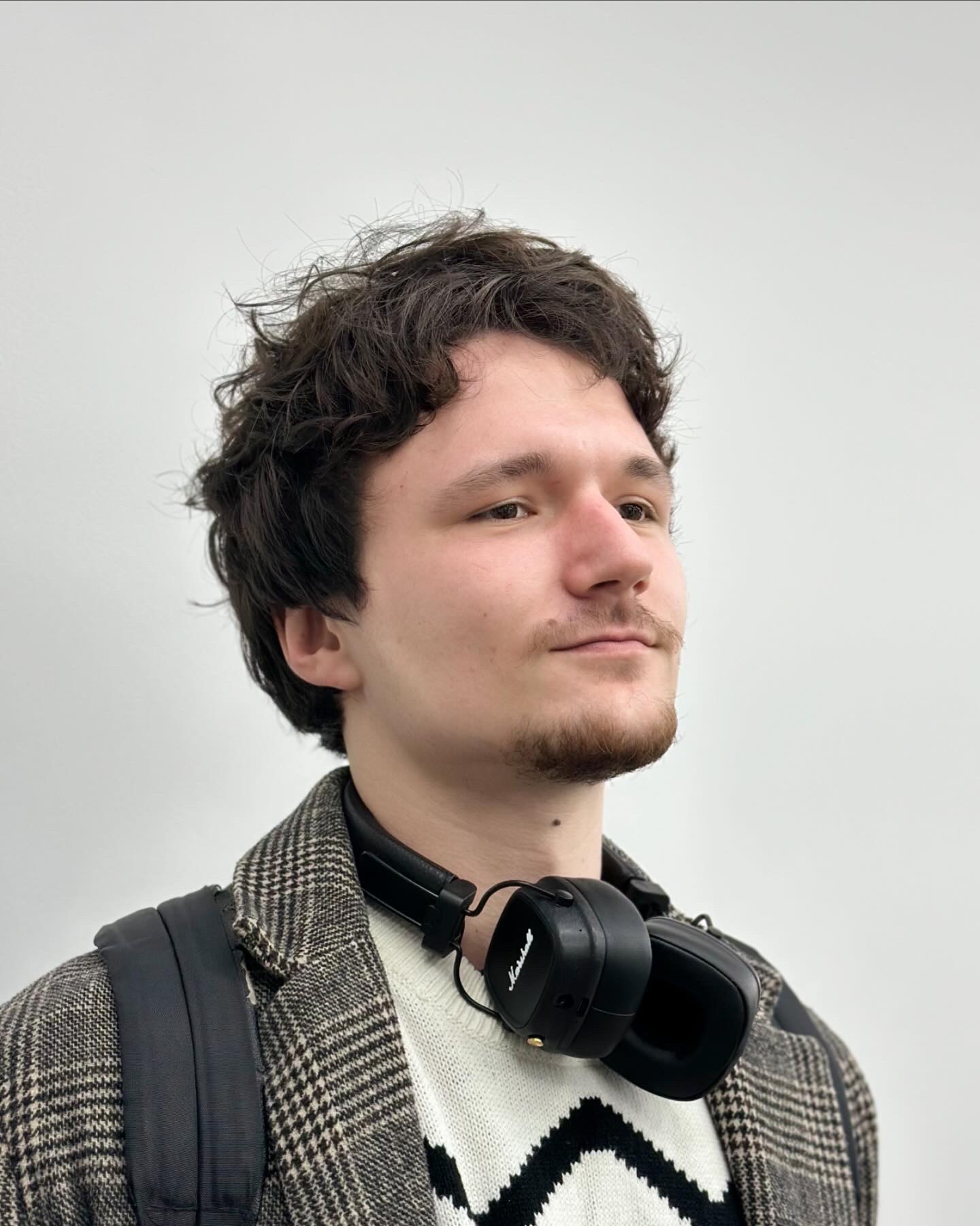 Młody mężczyzna z kręconymi włosami i delikatnym zarostem, po wizycie, spogląda w dal. Na szyi ma słuchawki Marshall, ubrany w sweter w zygzaki i marynarkę w pepitkę, z plecakiem na ramieniu.