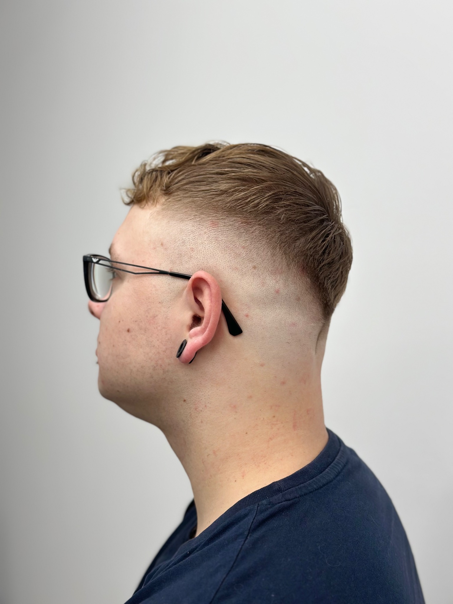 Męska fryzura z wycieniowanymi bokami i górą, widok z profilu, model w okularach i z tunelem w uchu na tle jasnej ściany.