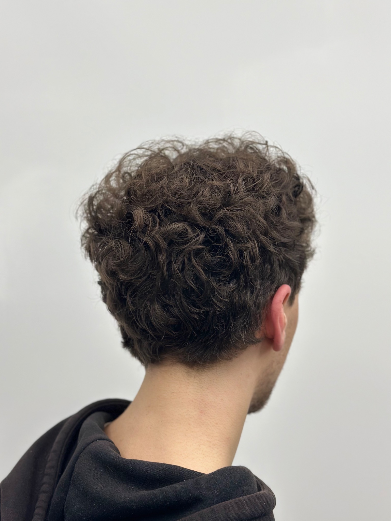 Tył głowy mężczyzny z krótkimi, kręconymi, brązowymi włosami na tle jasnej ściany, widoczny fragment czarnej bluzy z kapturem.