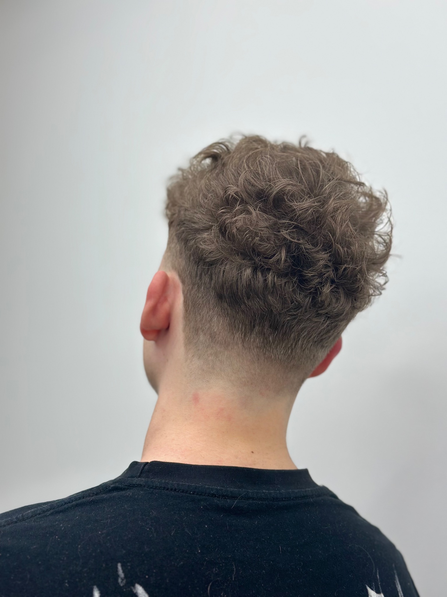 Krótkie, kręcone włosy mężczyzny po strzyżeniu, widok z tyłu, z delikatnym cieniowaniem na karku, na jednolitym, jasnym tle.