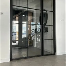 Nowoczesne, dwuskrzydłowe drzwi wewnętrzne z czarnymi szprosami i przeszkleniami, widok na wnętrze domu z rośliną doniczkową i krzesłem rattanowym.