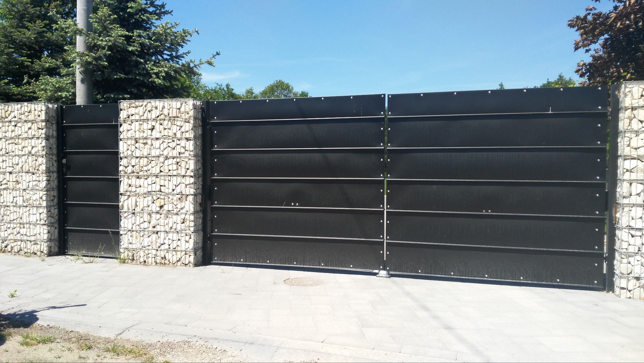 Nowoczesna brama wjazdowa z czarnymi panelami i gabionowymi słupkami wypełnionymi jasnymi kamieniami, widoczna na tle niebieskiego nieba i zieleni.