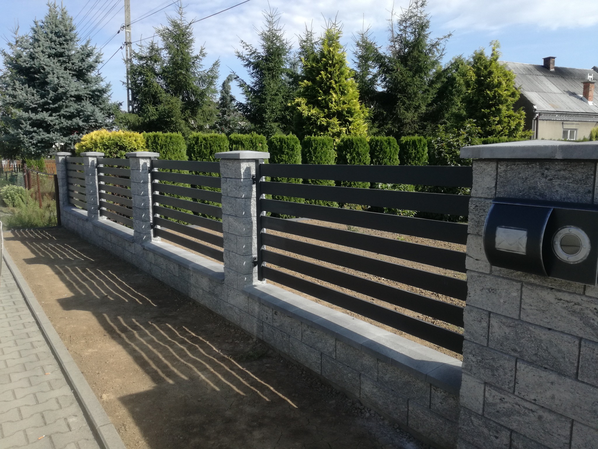 Nowoczesne ogrodzenie z poziomych, ciemnych paneli na kamiennej podmurówce i słupkach, z czarną skrzynką na listy umieszczoną na jednym ze słupków, w tle zielone drzewa i fragment domu.