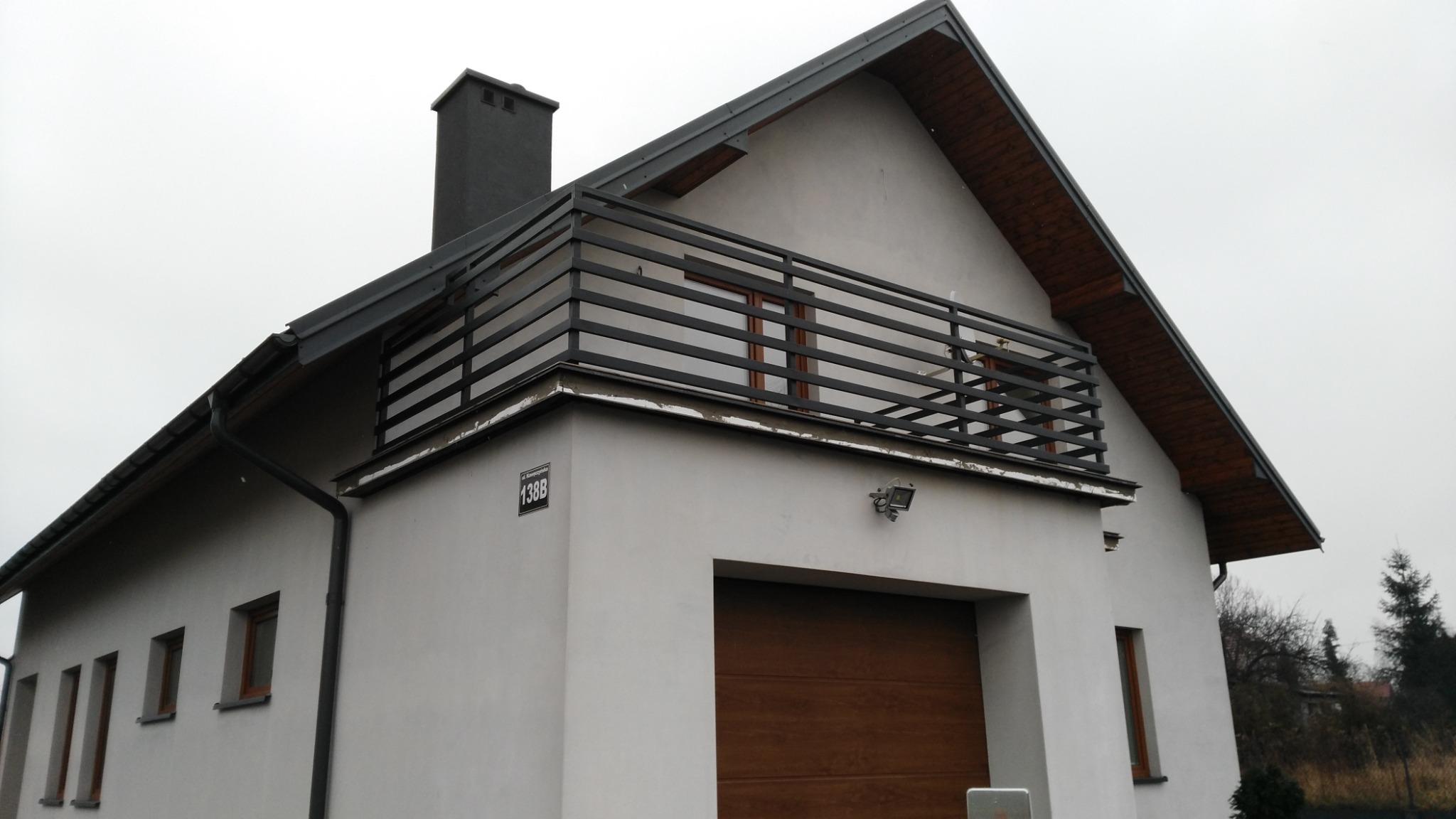 Nowoczesny dom z szarą elewacją, balkonem z czarną, poziomą balustradą i brązową bramą garażową.