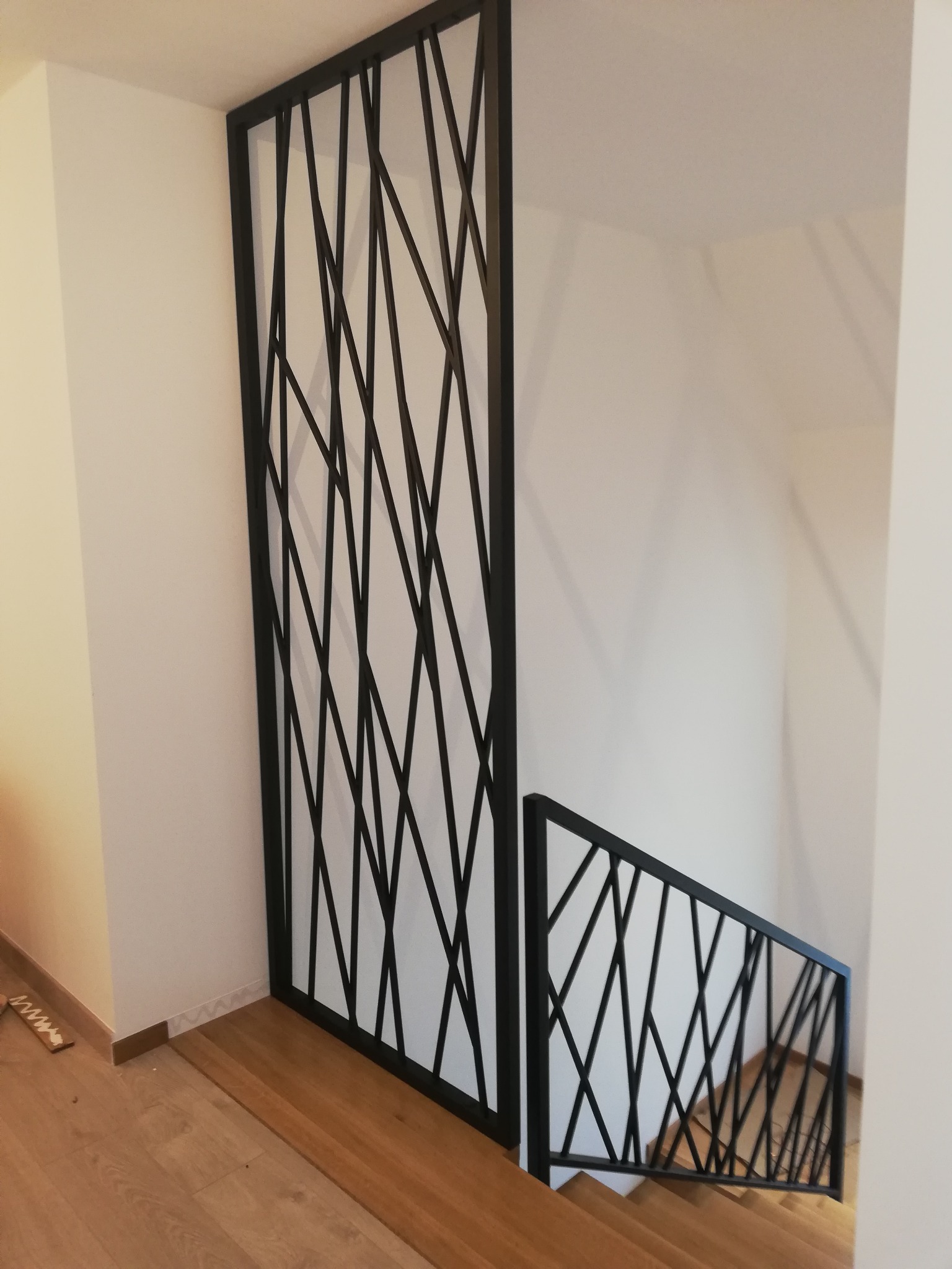 Nowoczesna, czarna metalowa balustrada schodowa z geometrycznym wzorem ukośnych linii, widoczna na tle białych ścian i drewnianych schodów.