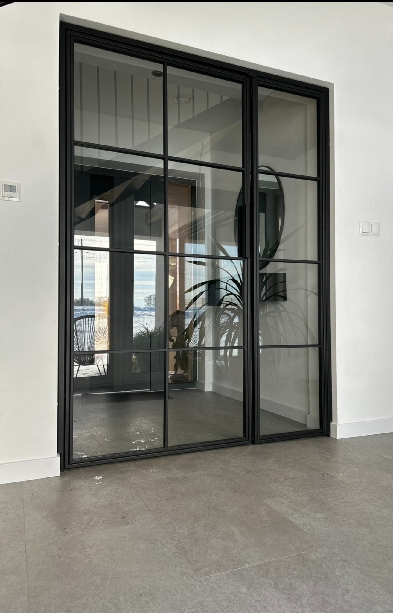 Nowoczesne, dwuskrzydłowe drzwi wewnętrzne z czarnymi szprosami i przeszkleniami, widok na wnętrze domu z rośliną doniczkową i krzesłem rattanowym.