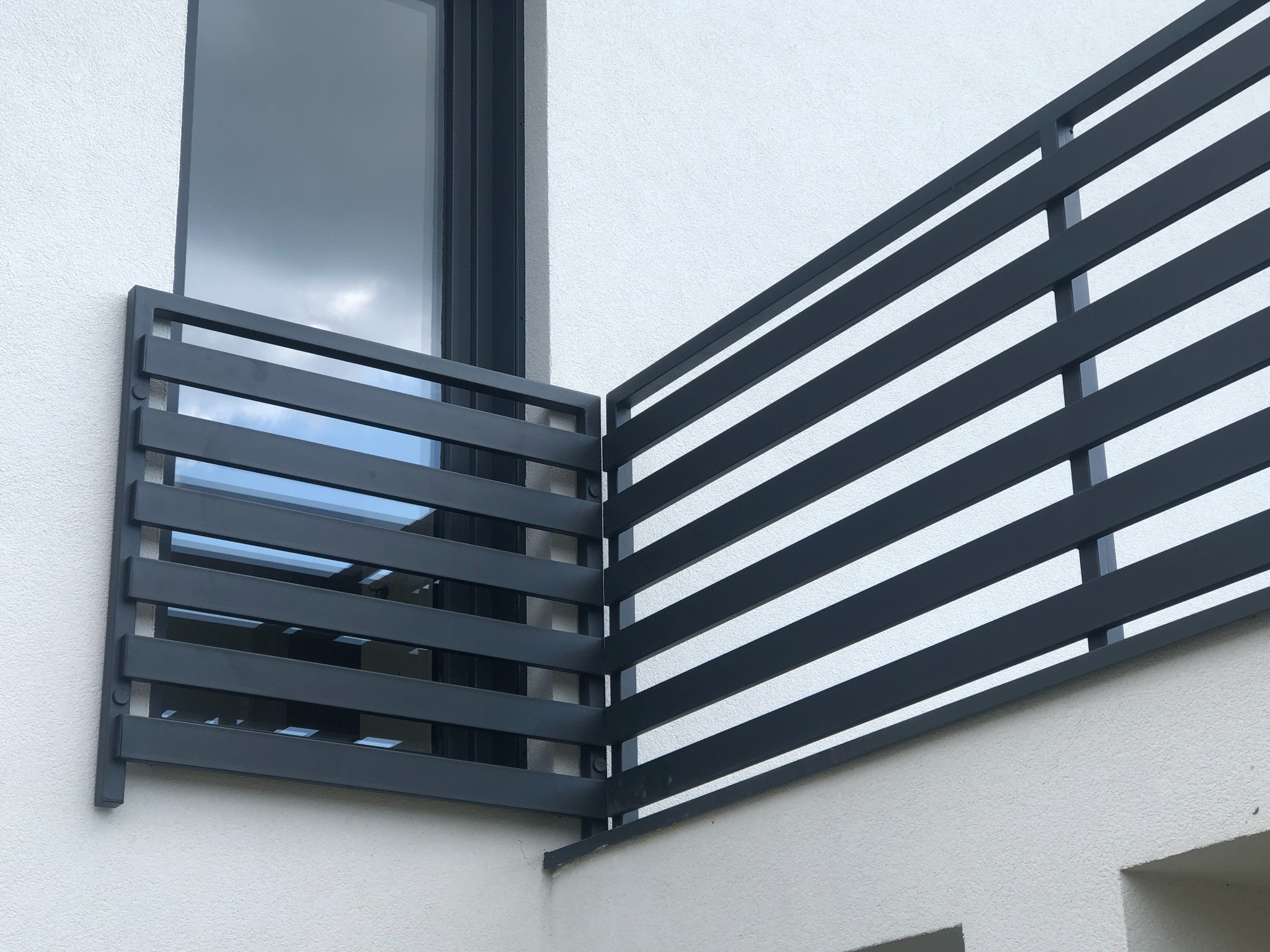 Nowoczesna, grafitowa balustrada balkonowa z poziomych profili, zamontowana przy oknie na białej elewacji budynku.