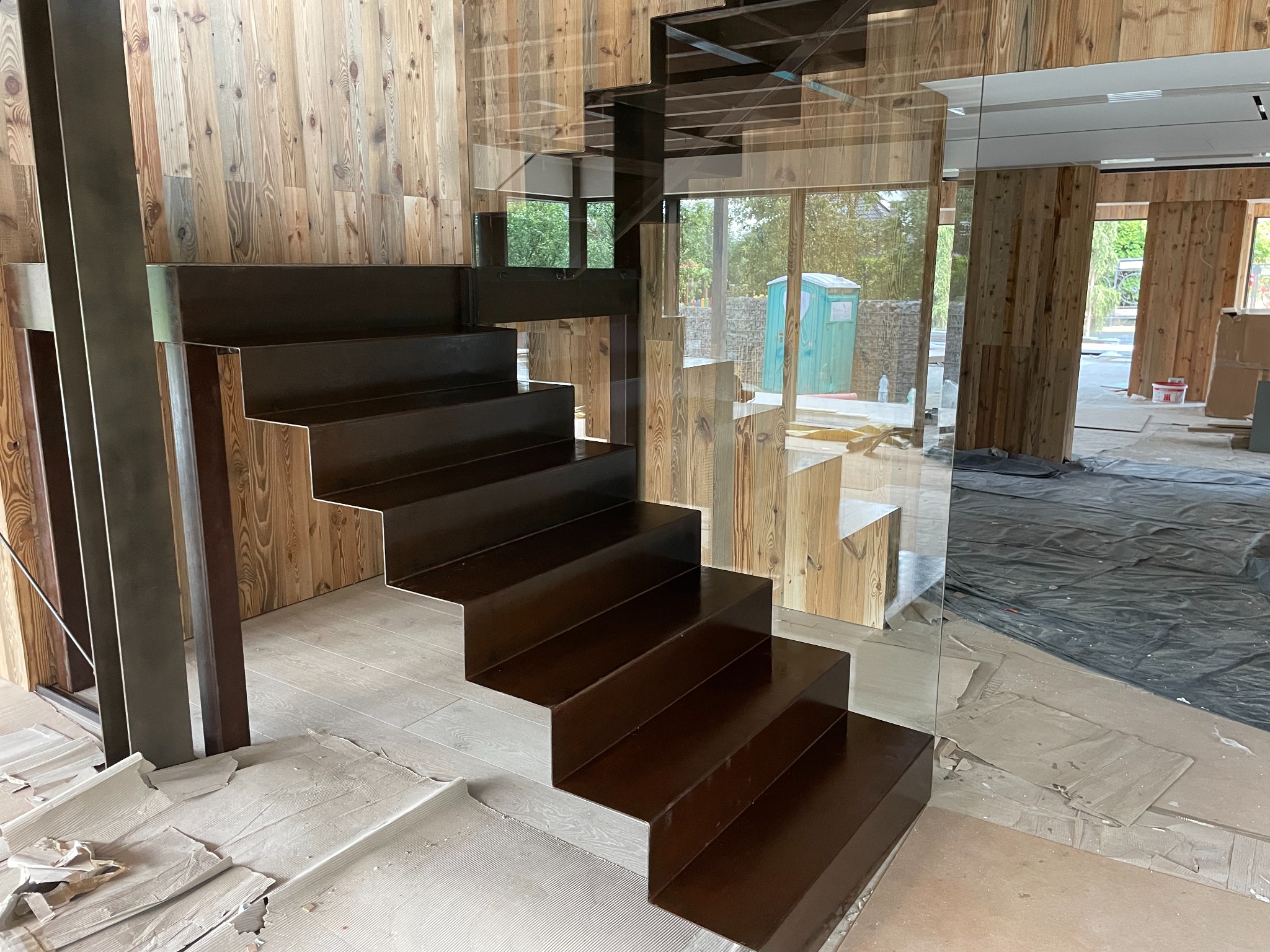 Nowoczesne schody z ciemnego metalu z szklaną balustradą w surowym wnętrzu z drewnianymi ścianami, zabezpieczone kartonem i folią malarską.