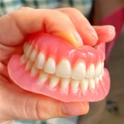 Ręka trzyma kompletną protezę zębową, widoczne detale akrylowej bazy i sztucznych zębów.