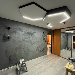 RavBud usługi ogólnobudowlane - Pokój po remoncie z szarą ścianą z efektem betonu, drewnianą podłogą i nowoczesnym oświetleniem sufitowym z białych paneli z wbudowanymi paskami LED, drabina malarska w rogu