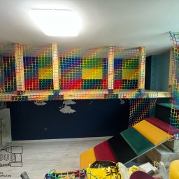 RavBud usługi ogólnobudowlane - Pokój dziecięcy z zabudową antresoli z kolorową siatką zabezpieczającą i wejściem po schodkach z kolorowymi poduszkami, widoczny fragment ściany z malunkami chmur i logo firmy remontowej w rogu.