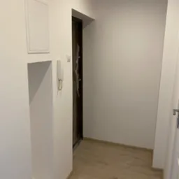 Nowo wykończony korytarz z drewnopodobną podłogą, białe ściany z wnęką i panelem dostępowym, drzwi wejściowe zabezpieczone folią ochronną.