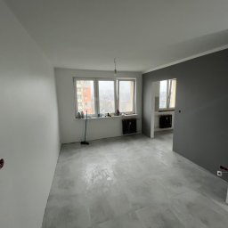 RavBud usługi ogólnobudowlane - Pusty pokój po remoncie z szarą ścianą, jasnymi płytkami podłogowymi i odsłoniętymi gniazdkami elektrycznymi.