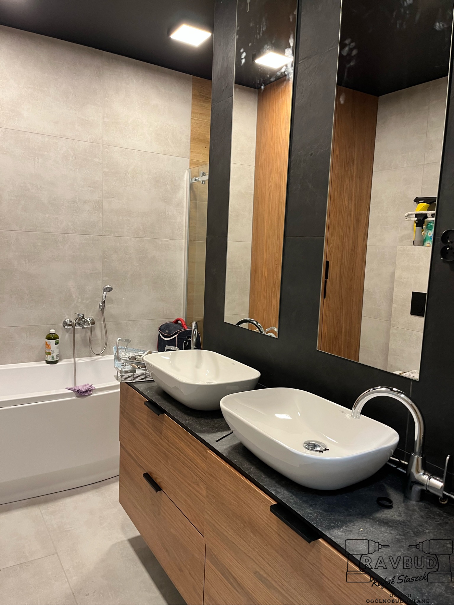 Nowoczesna łazienka z dwiema owalnymi umywalkami nablatowymi, drewnianą szafką pod umywalkę z czarnym blatem, dużymi lustrami w czarnych ramach, beżowymi płytkami na ścianach i wanną po lewej stronie.