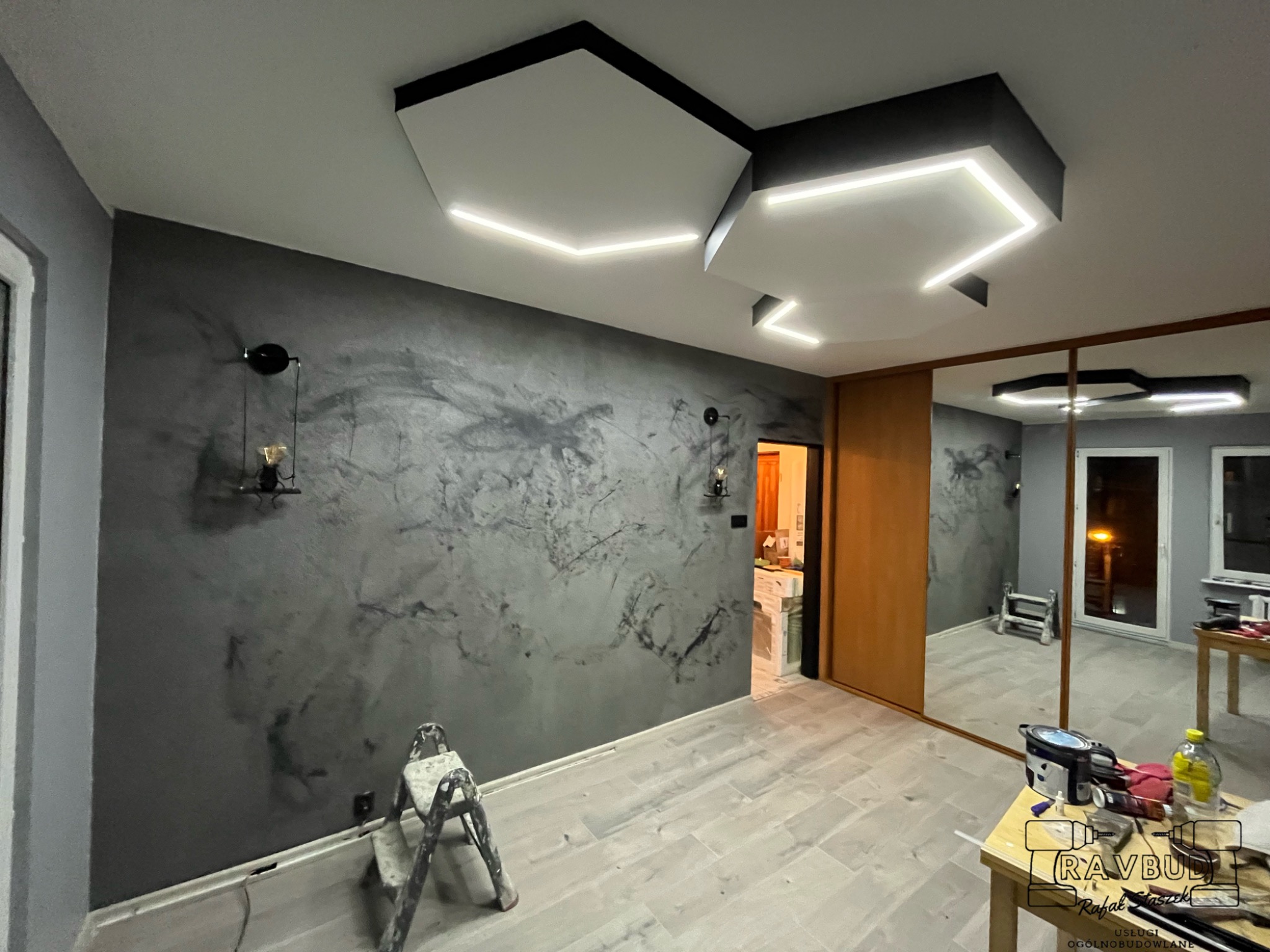 Pokój po remoncie z szarą ścianą z efektem betonu, drewnianą podłogą i nowoczesnym oświetleniem sufitowym z białych paneli z wbudowanymi paskami LED, drabina malarska w rogu