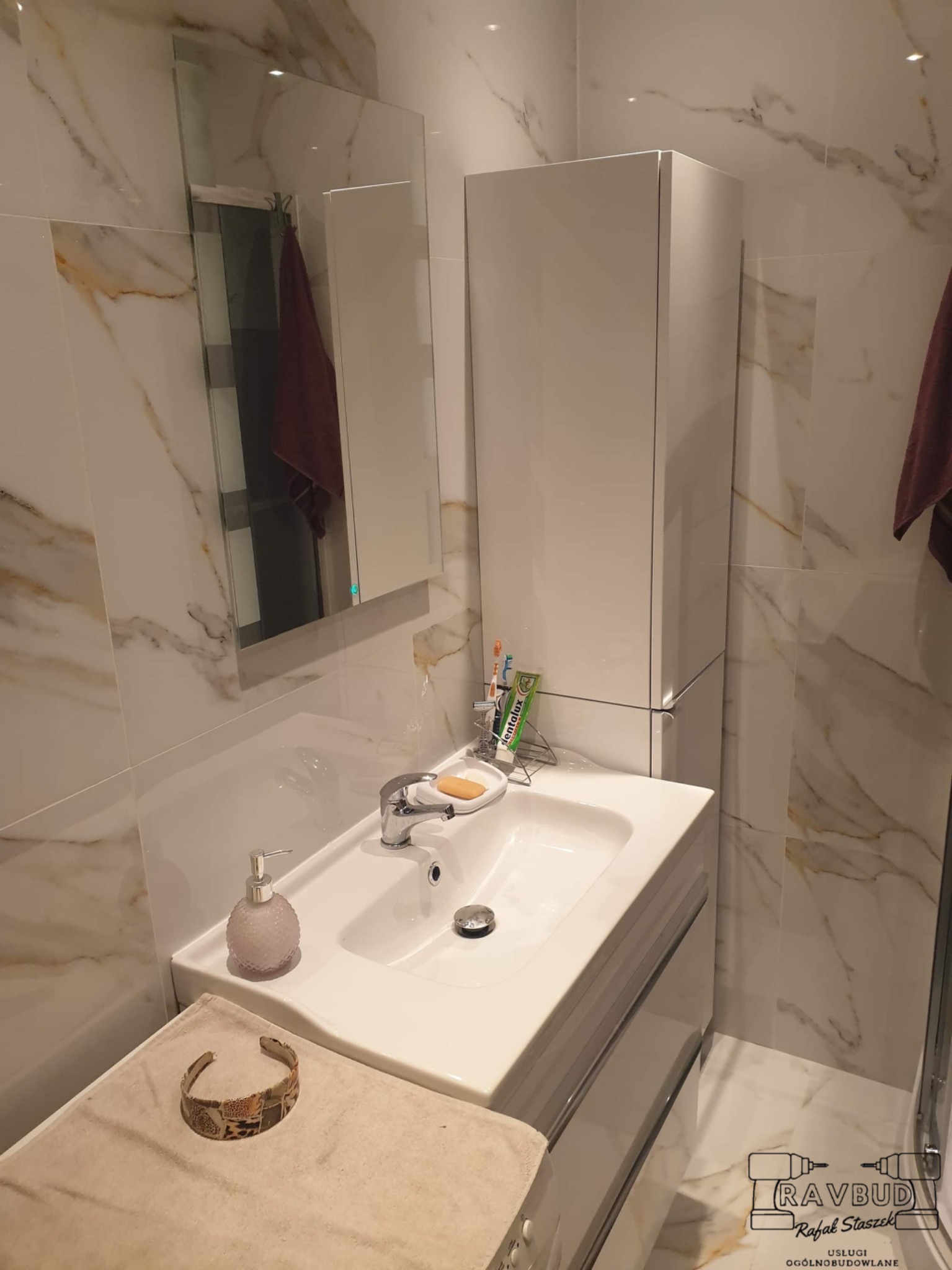 Umywalka z szafką w nowoczesnej łazience wykończonej płytkami imitującymi marmur, widoczny ręcznik, mydło i akcesoria do higieny jamy ustnej, na blacie leży ozdobna bransoletka.