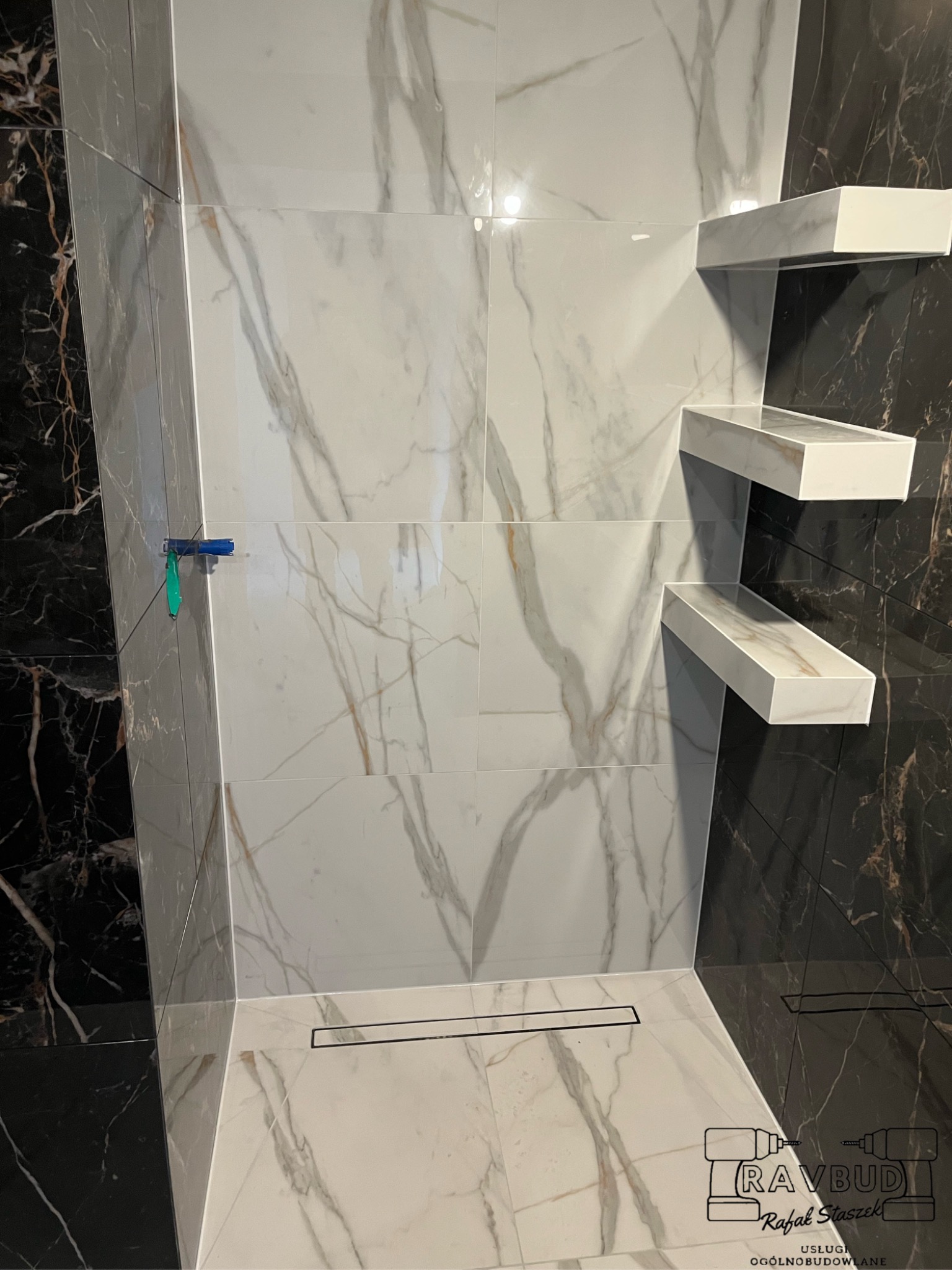 Wykończona kabina prysznicowa z białymi i czarnymi płytkami imitującymi marmur, z wbudowanymi półkami i odpływem liniowym, zabezpieczona folią w narożniku.