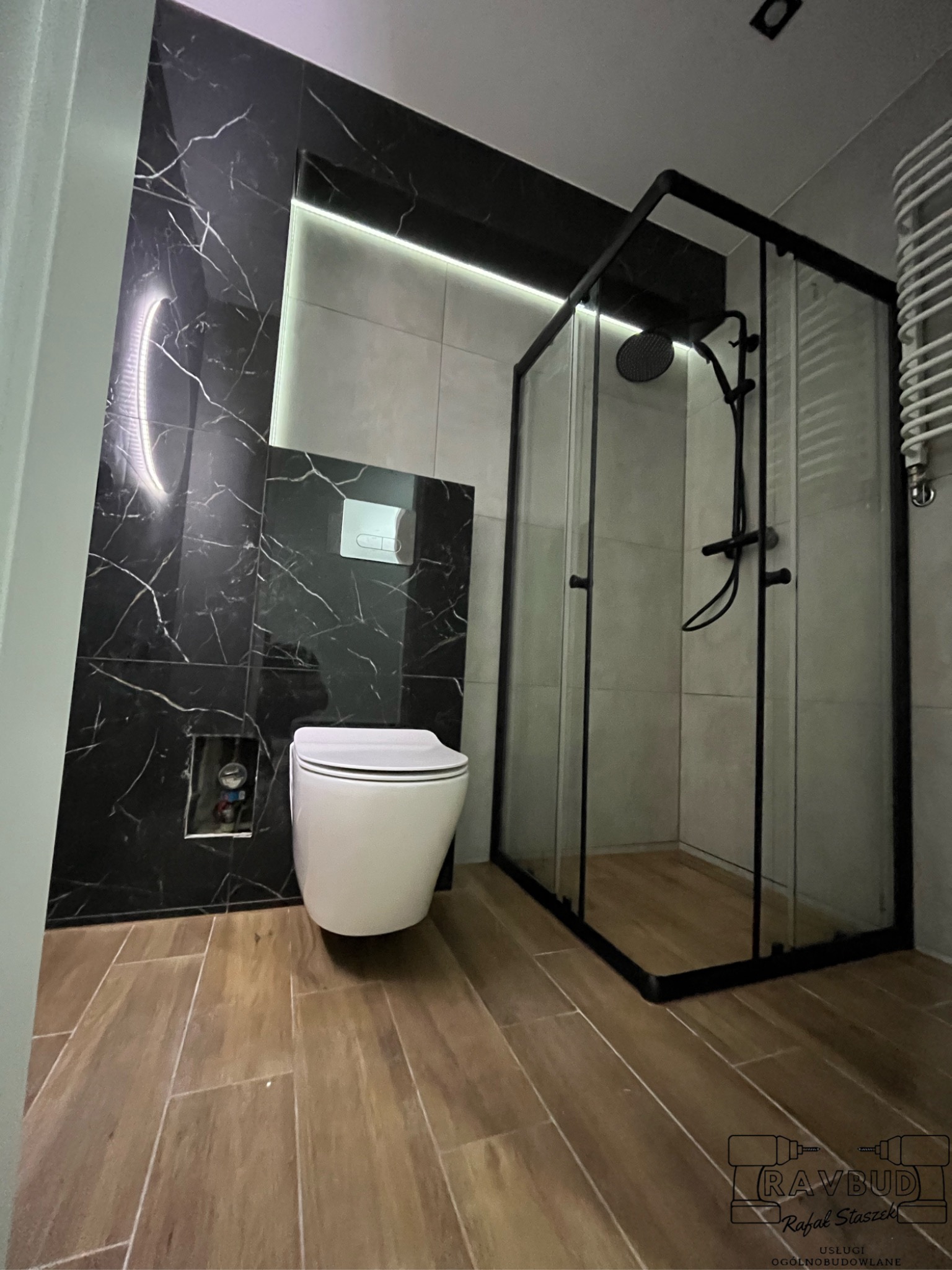 Nowoczesna łazienka z czarnymi płytkami imitującymi marmur, podświetleniem LED, kabiną prysznicową z czarną ramą i drewnopodobną podłogą. Uwagę zwraca wisząca miska WC i minimalistyczny design.