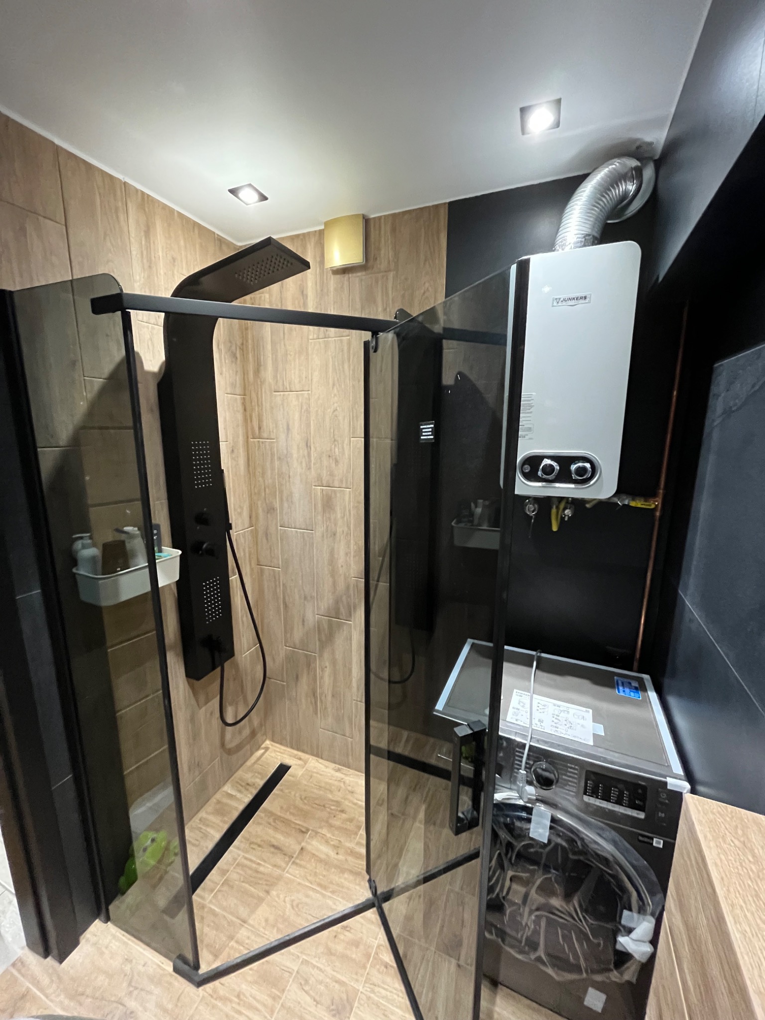 Nowoczesna kabina prysznicowa typu walk-in z czarnymi profilami i deszczownicą, umieszczona w łazience z płytkami imitującymi drewno, obok pralka w folii i piec gazowy Junkers.