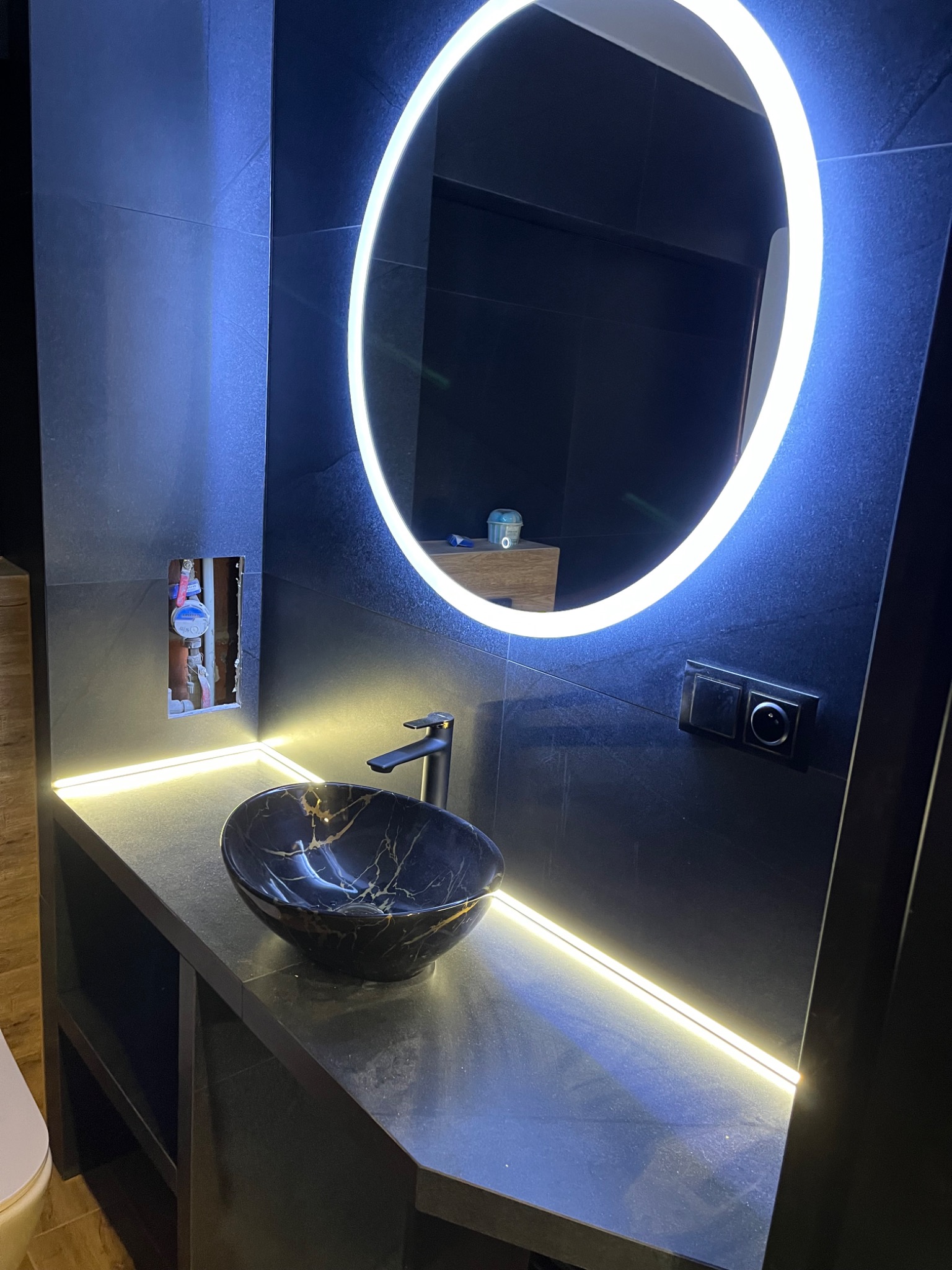 Nowoczesna łazienka z czarnymi płytkami, okrągłym lustrem z podświetleniem LED, umywalką z czarnego marmuru i widocznymi instalacjami hydraulicznymi w ścianie.