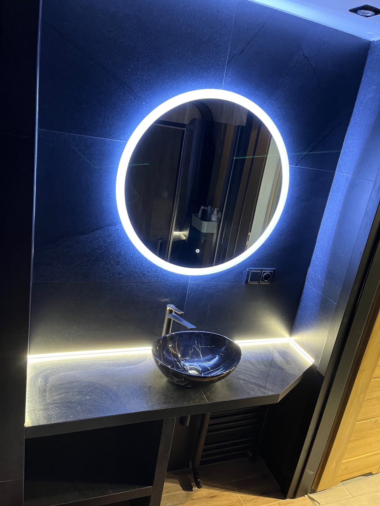 Nowoczesna łazienka z czarnymi płytkami, okrągłym lustrem z podświetleniem LED i umywalką nablatową w kolorze granatowym z marmurkowym wzorem.