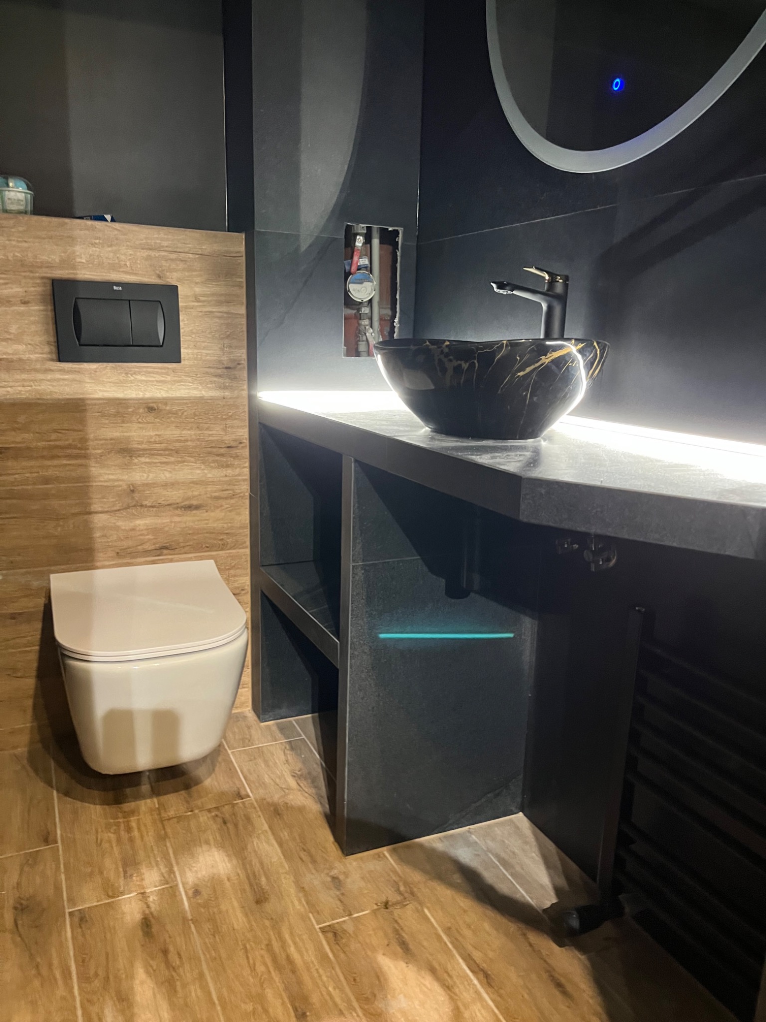 Nowoczesna łazienka z wiszącą toaletą, drewnopodobnymi płytkami na podłodze i ścianie, czarną umywalką w marmurkowy wzór na czarnym blacie z podświetleniem LED oraz okrągłym lustrem z podświetleniem.