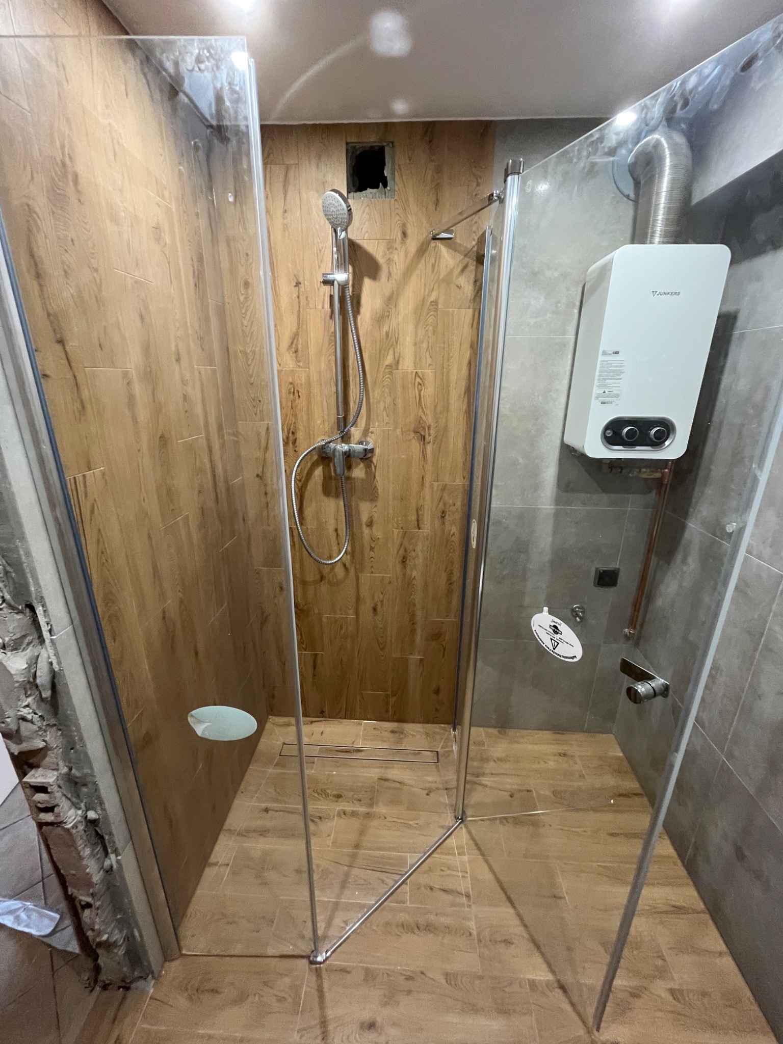 Wnętrze kabiny prysznicowej z drewnopodobnymi płytkami, widoczny bojler Junkers na szarej ścianie oraz niedokończone wykończenie ściany obok kabiny.