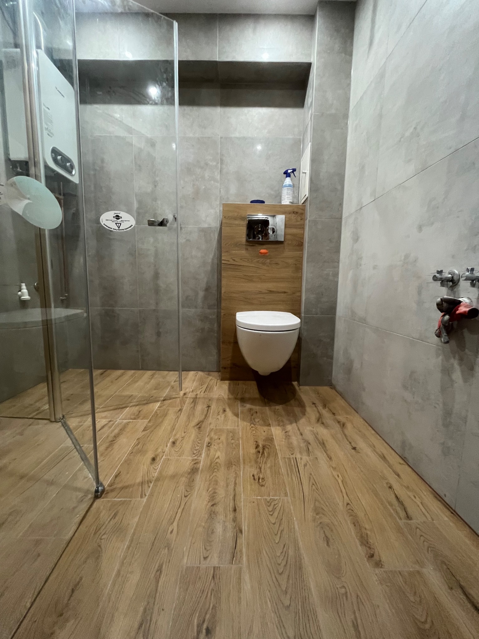 Nowoczesna łazienka z drewnopodobną podłogą, szarą glazurą, kabiną prysznicową ze szkła i wiszącą toaletą z zabudową z drewnianych paneli.