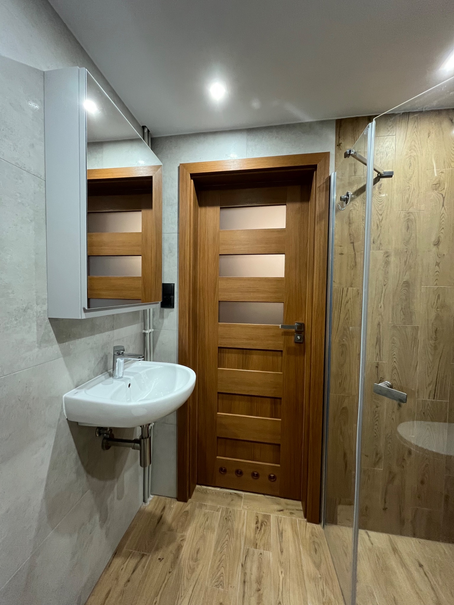 Nowoczesna łazienka z drewnianymi akcentami: umywalka nablatowa z szafką lustrzaną, drewniane drzwi z przeszkleniami i kabina prysznicowa z panelami imitującymi drewno.