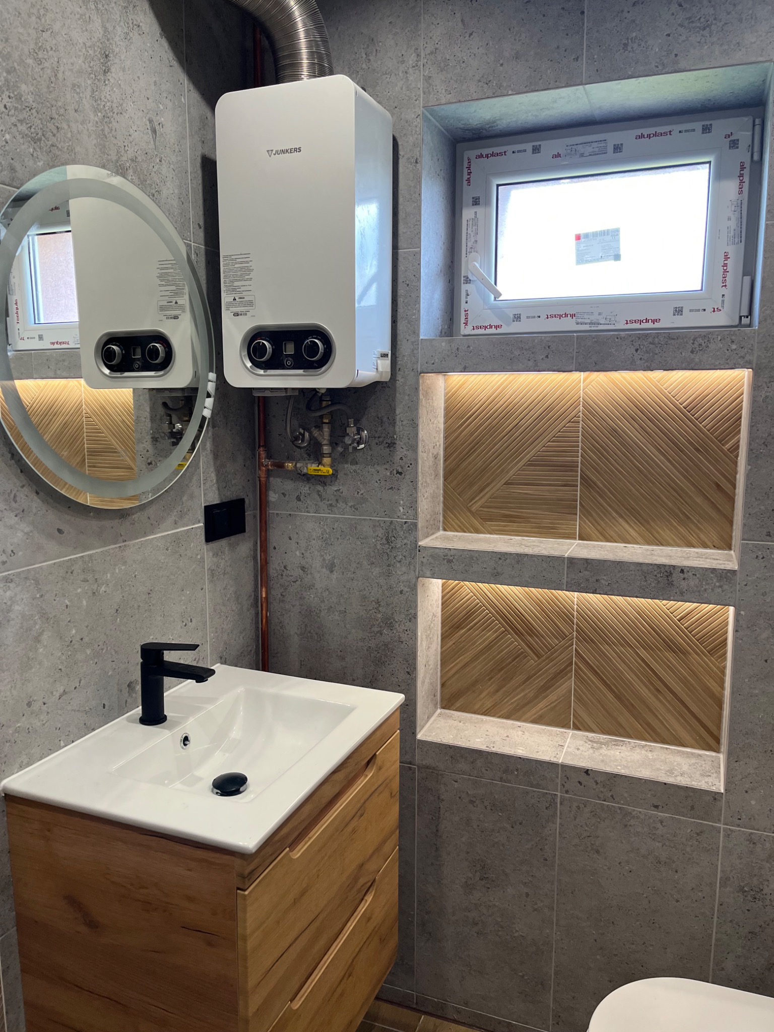 Nowoczesna łazienka z szarymi płytkami, drewnianą szafką pod umywalką z czarną baterią, okrągłym lustrem, białym bojlerem Junkers i wnękami z dekoracyjnym drewnem.