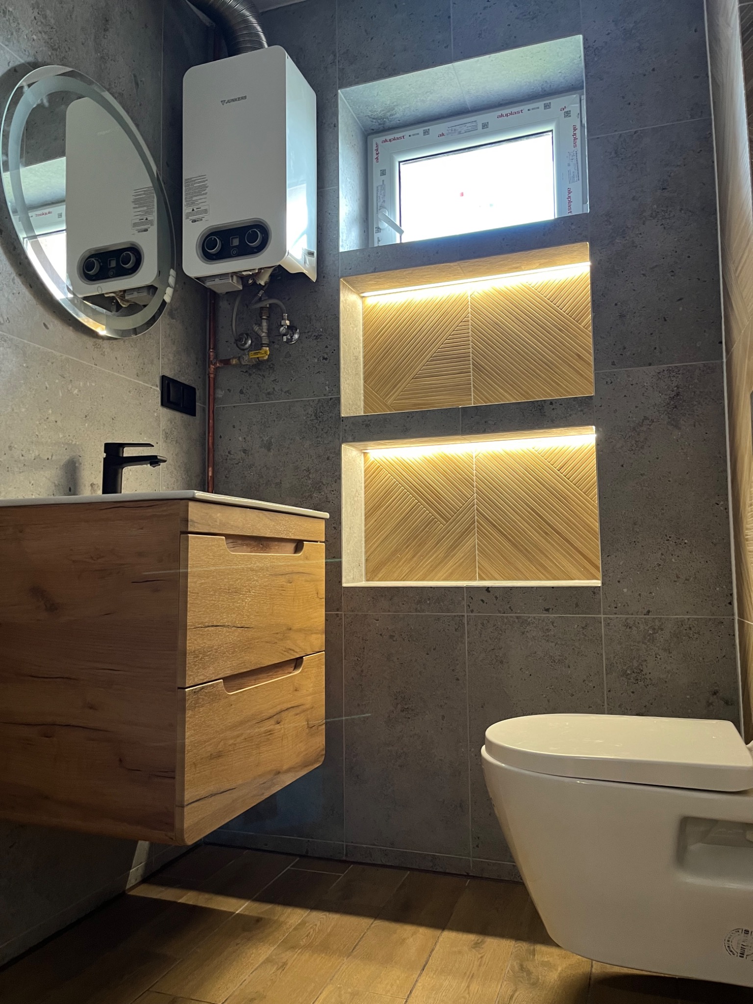 Nowoczesna aranżacja wnętrza łazienki z szarą, kamienną okładziną ścian, podwieszaną szafką z drewnianym fornirem, owalnym lustrem z podświetleniem LED, wiszącą miską WC i dwoma podświetlanymi...