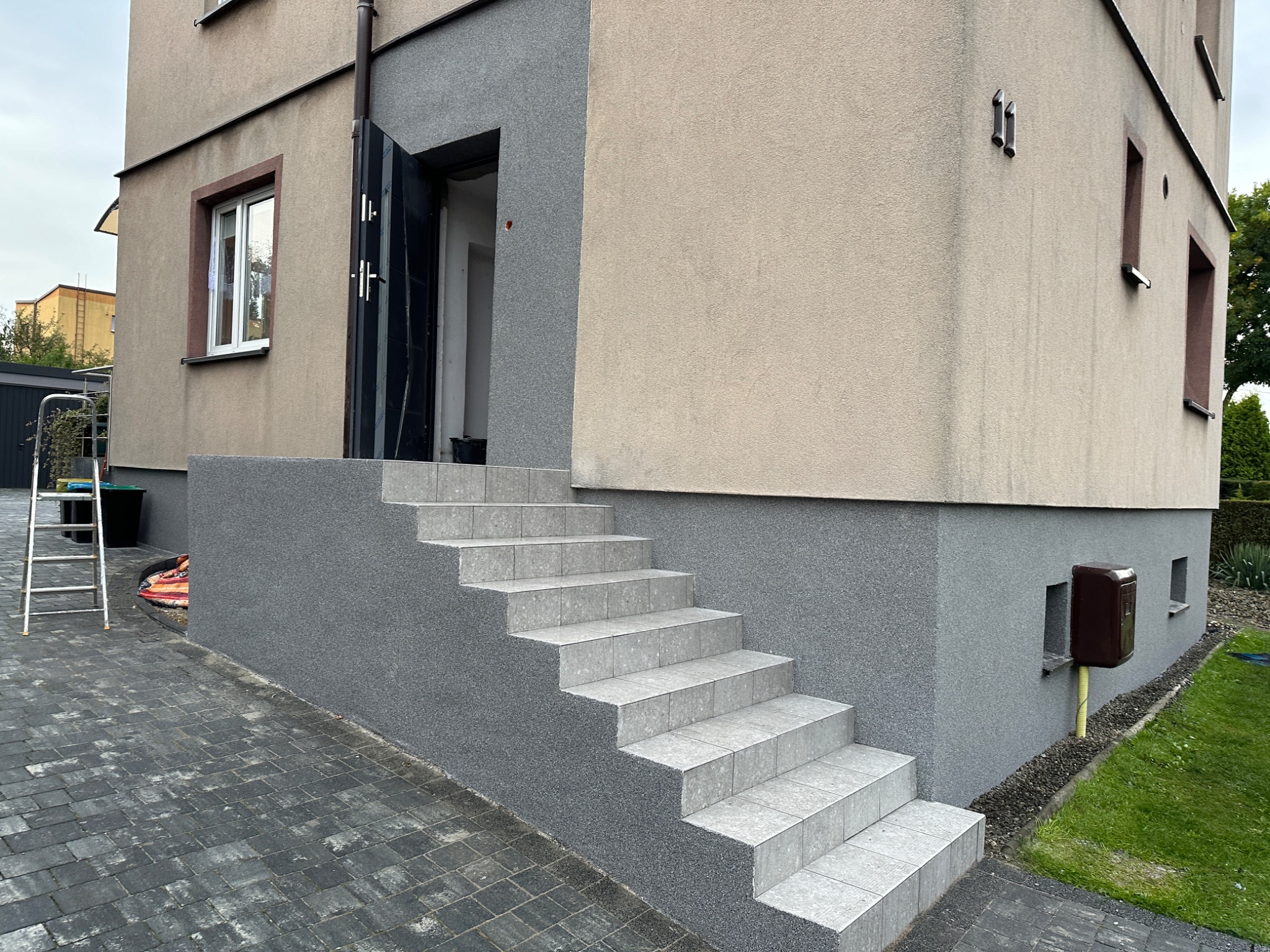 Wykończone płytkami schody wejściowe do domu w Katowicach, z szarą elewacją i widocznym fragmentem chodnika z kostki brukowej.