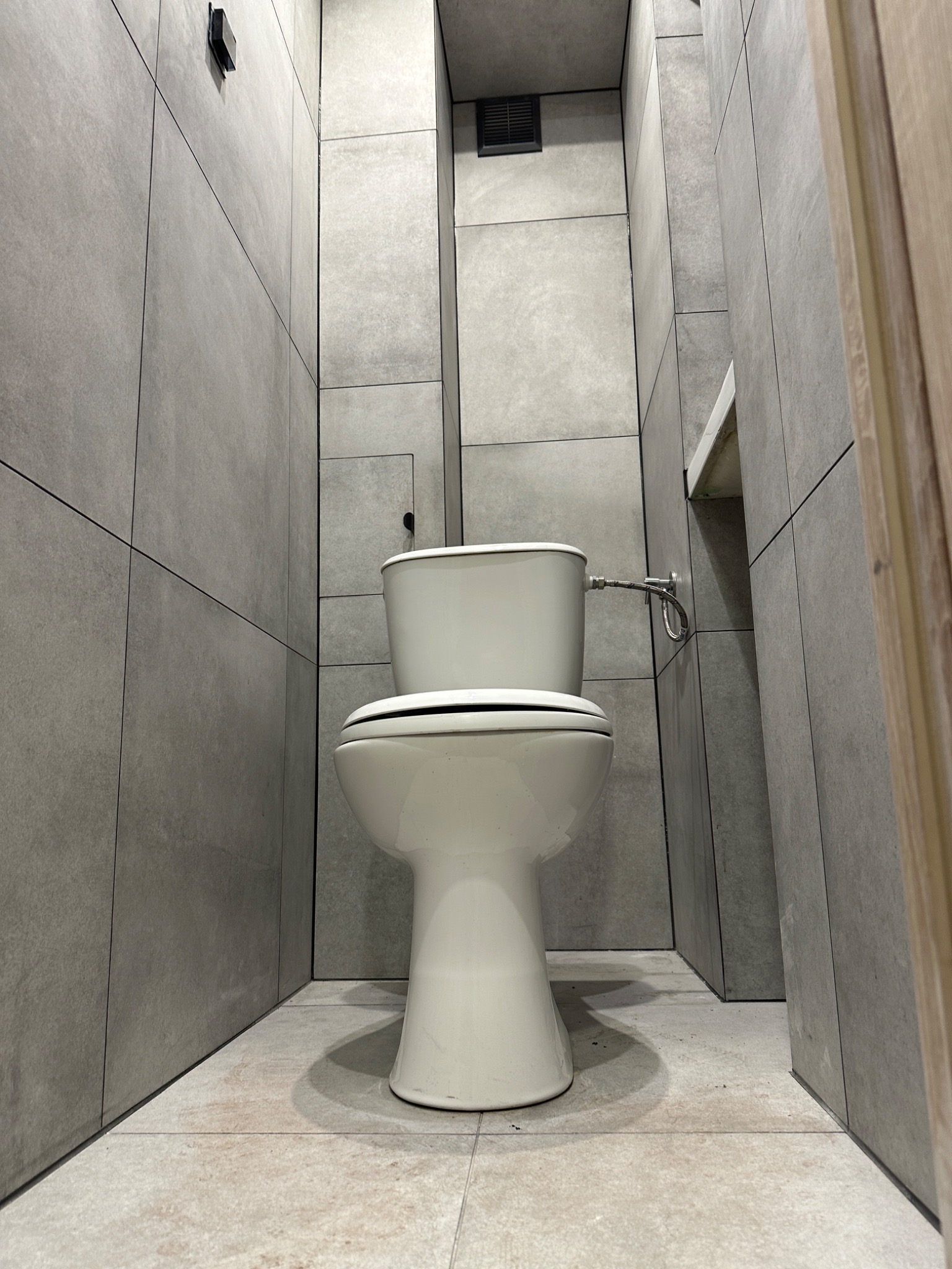 Wąska toaleta z szarymi płytkami wielkoformatowymi i białą ceramiką sanitarną, widok z dołu podkreślający pionowe linie i minimalistyczny design, z fragmentem drewnianej ościeżnicy po prawej.