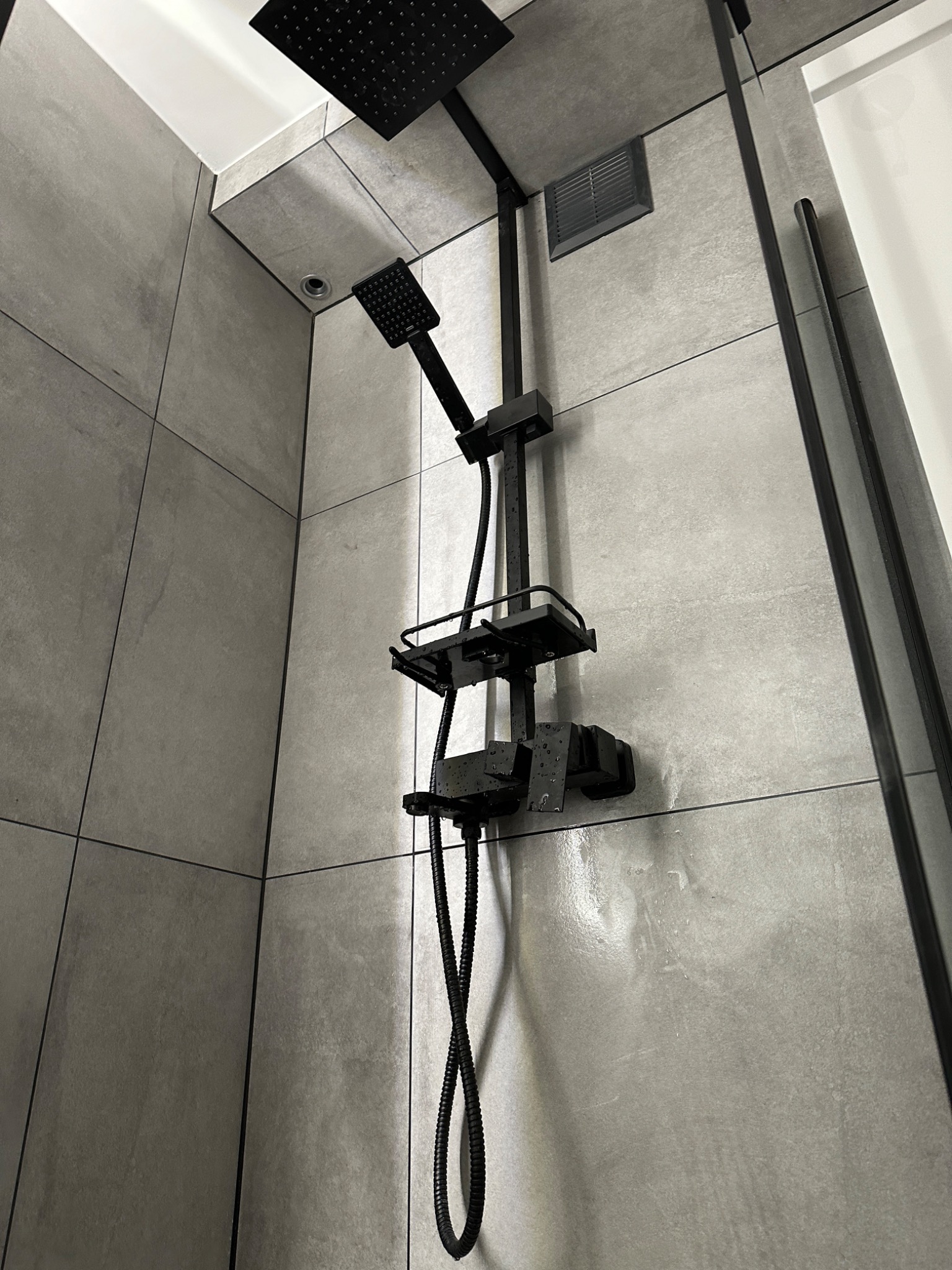 Nowoczesny panel prysznicowy w kolorze czarnym, zamontowany na ścianie wyłożonej szarymi płytkami w łazience, widoczne krople wody po użyciu.