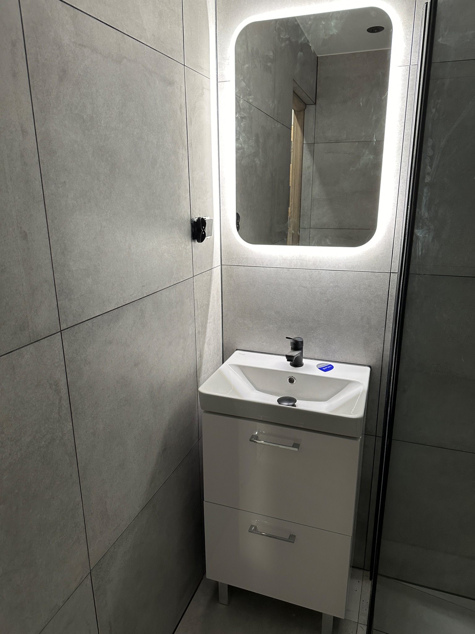 Nowoczesna umywalka z szafką w szarej łazience, podświetlane lustro LED, minimalistyczny design, czarna bateria, widoczne płytki wielkoformatowe.