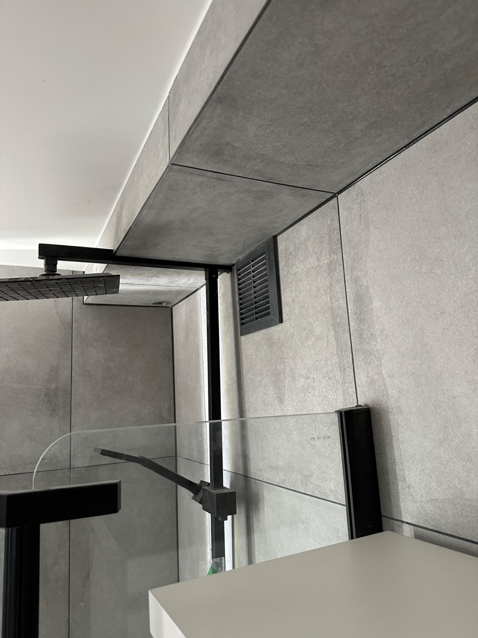 Nowoczesna kabina prysznicowa z szarą, kamienną okładziną, czarną armaturą i szklaną ścianką działową, widoczny fragment wentylacji.