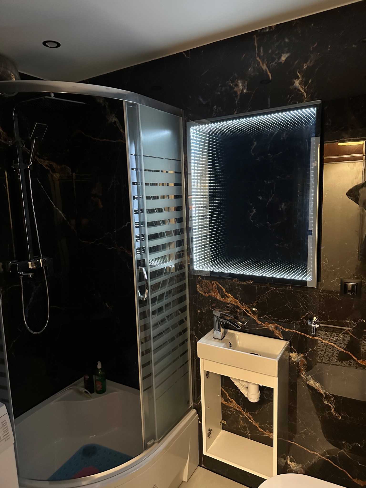 Nowoczesna łazienka z kabiną prysznicową narożną, umywalką wiszącą na tle ściany z czarnego marmuru i lustrem nieskończoności z podświetleniem LED.