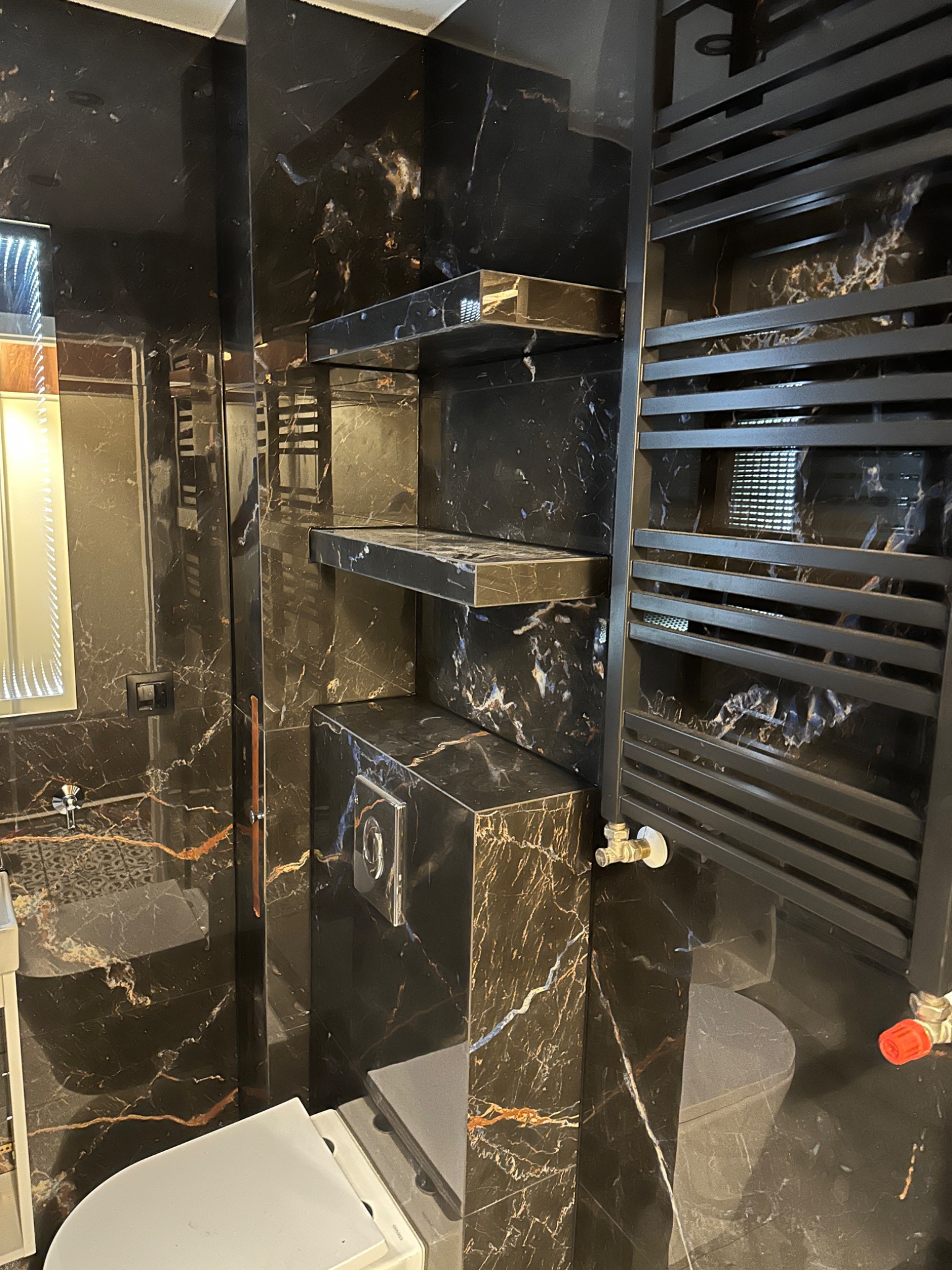 Nowoczesna łazienka z czarnymi marmurowymi ścianami, półkami i grzejnikiem drabinkowym, podświetlane lustro i wisząca toaleta.