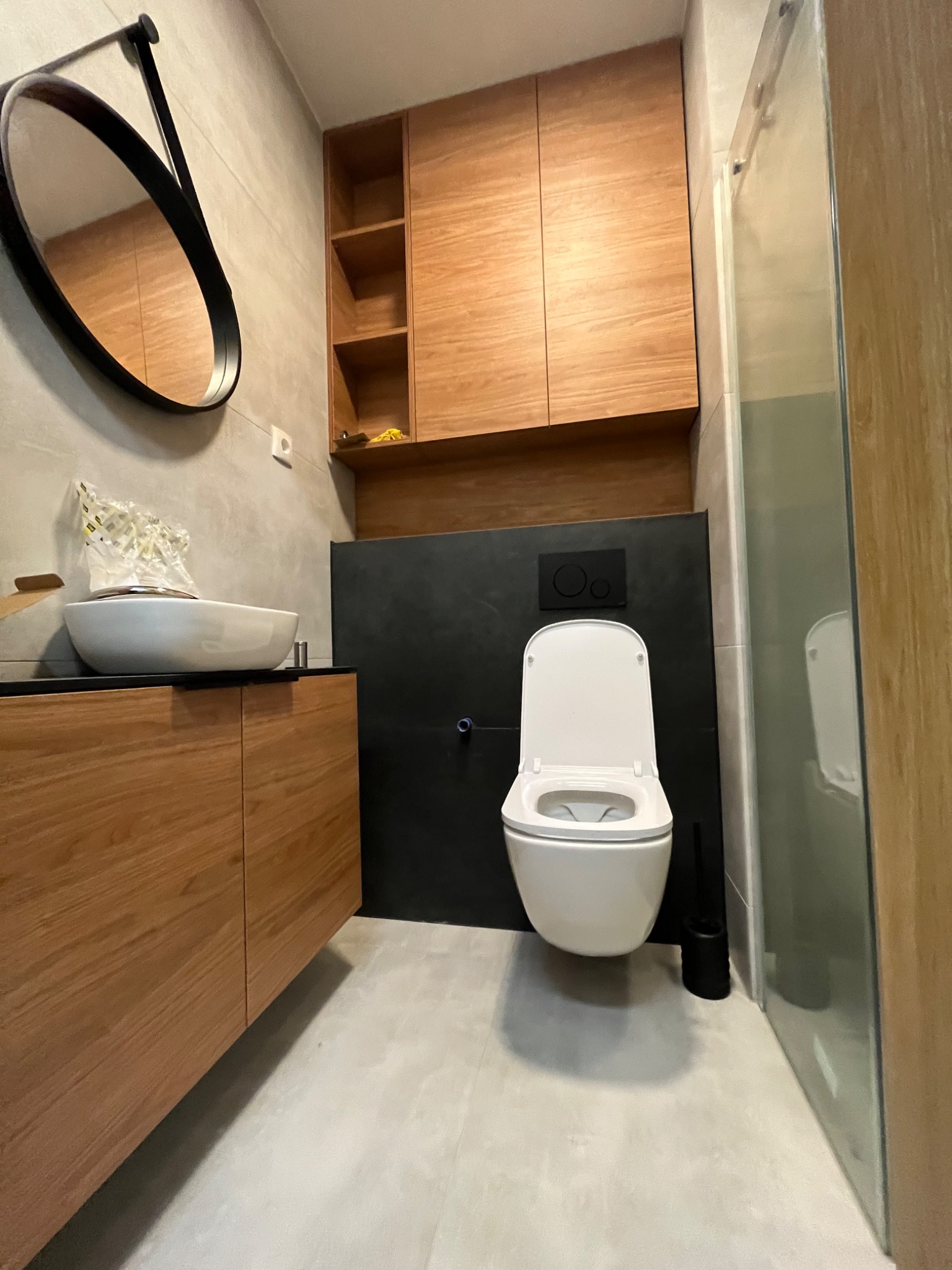 Nowoczesna łazienka z drewnianymi szafkami, owalnym lustrem w czarnej ramie i białą umywalką nablatową, wisząca toaleta z czarnym panelem spłukującym i kabina prysznicowa ze szklanymi drzwiami.