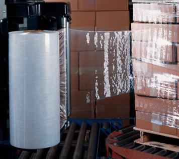 Rolka przezroczystej folii stretch na tle częściowo owiniętych kartonowych pudeł na linii produkcyjnej.