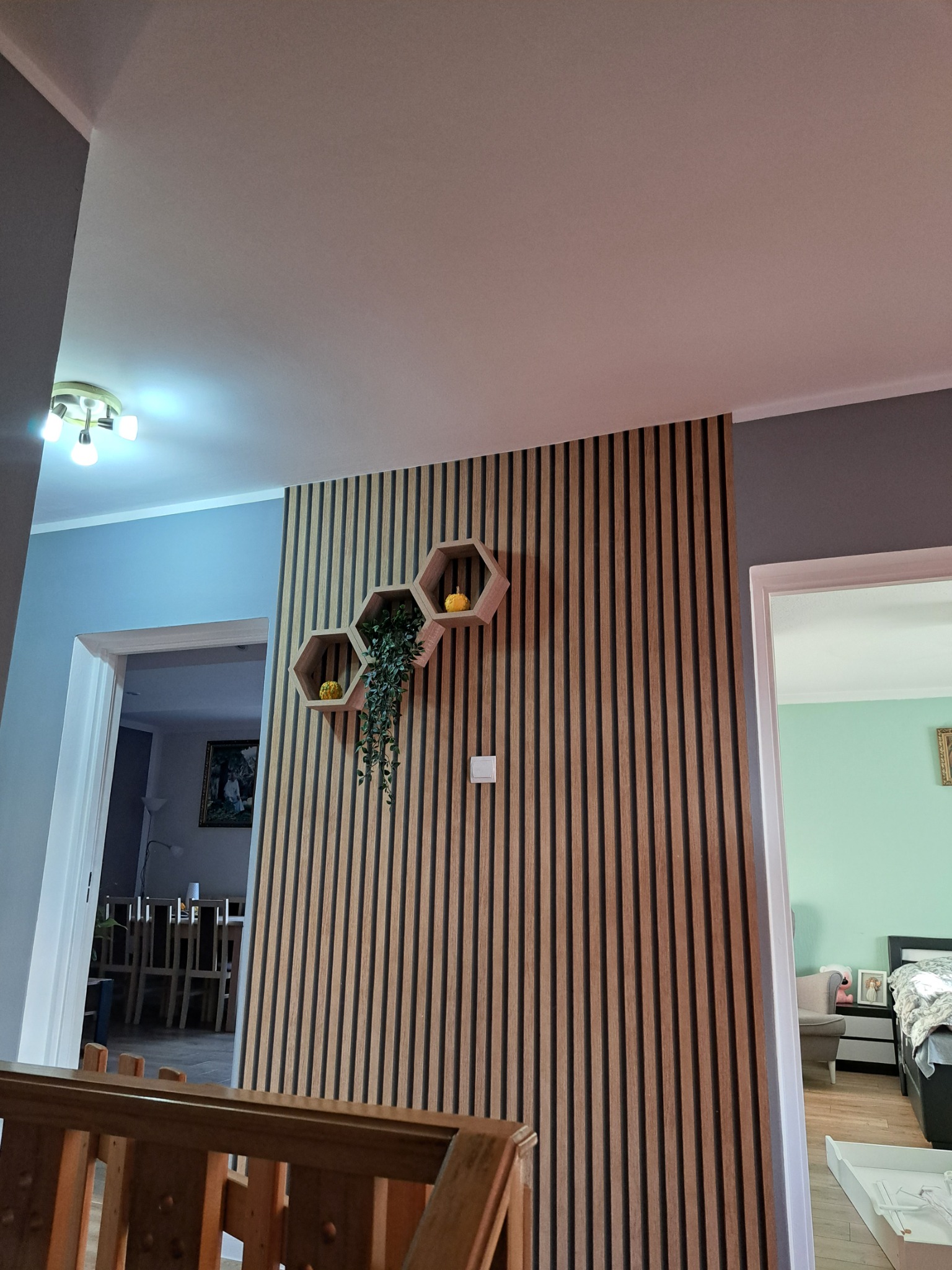 Drewniana ściana z pionowych paneli dekoracyjnych, z trzema sześciokątnymi półkami, na których znajdują się roślina i dekoracyjne dynie, widok na pokój przez otwarte drzwi.