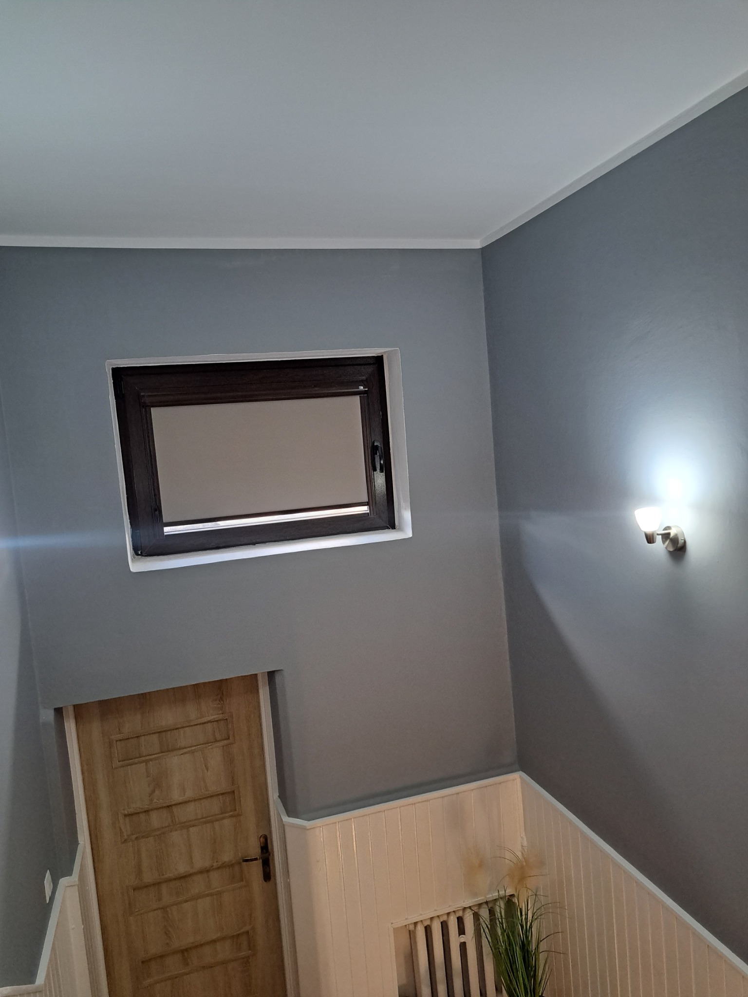 Szare wnętrze po remoncie: gładkie ściany, biały sufit, drewniane drzwi i okno z roletą, oświetlenie ścienne, dekoracyjny grzejnik z rośliną.