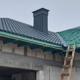 Ciemnoszary dach z blachodachówki z kominem w trakcie budowy, widoczna surowa ściana budynku z betonu komórkowego i drewniana drabina oparta o ścianę.