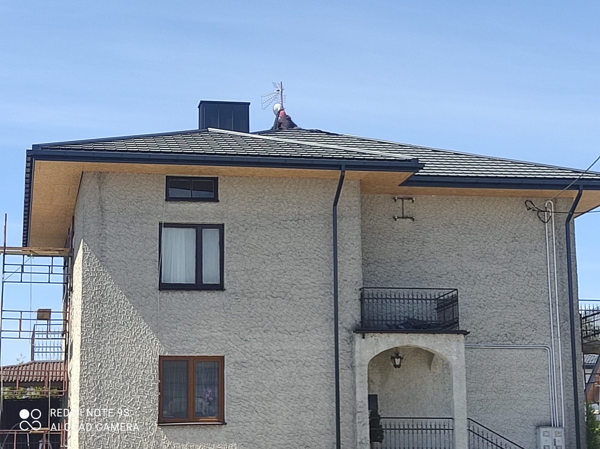 Dwupiętrowy dom z nowym, ciemnym dachem z dachówki bitumicznej; na dachu widoczna antena i osoba pracująca, z boku rusztowanie.