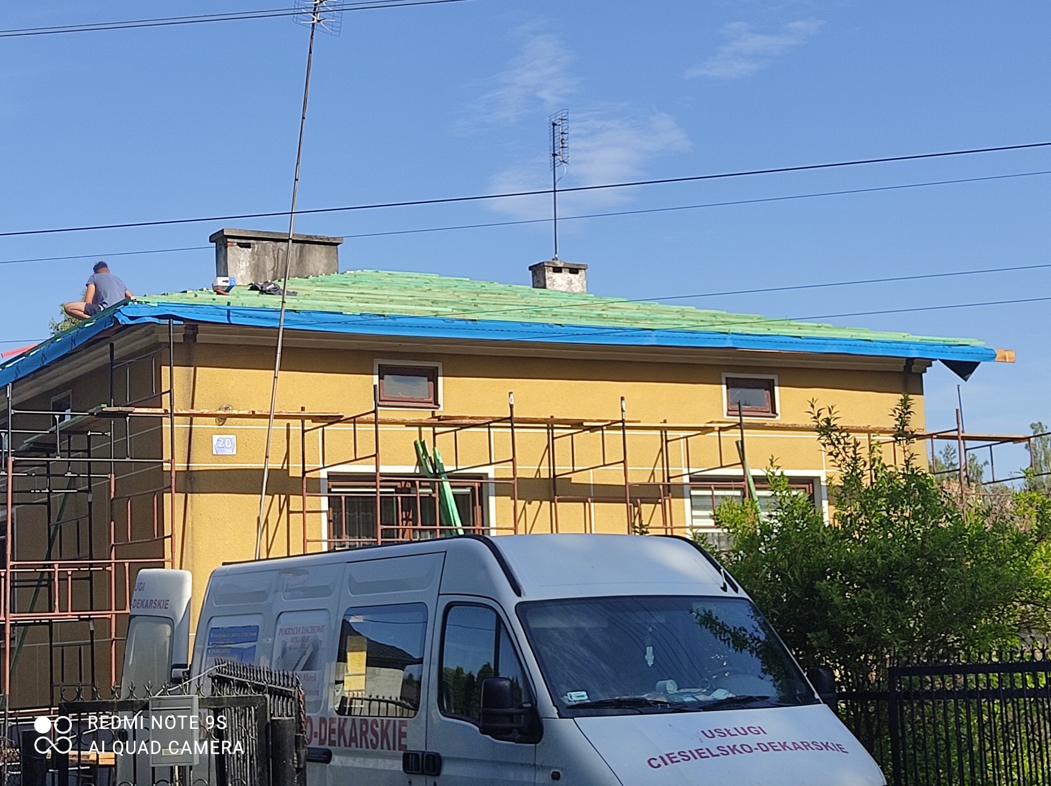 Pracownik siedzi na dachu budynku z żółtą elewacją, zabezpieczonym niebieską folią, widoczne rusztowanie i firmowy bus z napisem 'Usługi ciesielsko-dekarskie'.