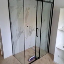 Nowoczesna kabina prysznicowa z czarnymi profilami i marmurowymi płytkami na ścianach, częściowo widoczny regał z narzędziami w tle, na podłodze leży przyssawka do płytek.