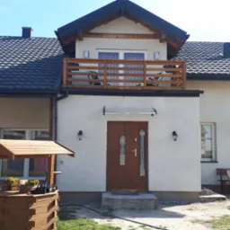 Nowoczesny dom jednorodzinny z balkonem, ciemnym dachem i studnią z kwiatami przed wejściem.